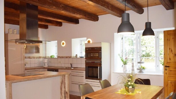 A modern kitchen with wooden beams, a dining area with a wooden table, and a large window with plants. - "Experience Tranquility and Adventure in Ole Hof Risum-Lindholm!" (Risum-Lindholm)