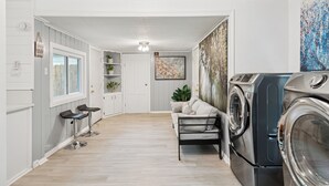 Un lavadero con lavadora, secadora y un banco. Se ve una puerta que da a otra habitación con una ventana y una planta.