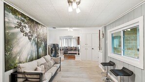 Una sala de estar moderna con un gran mural de un bosque, un sofá, una mesa de centro y una zona de cocina con un ventanal.