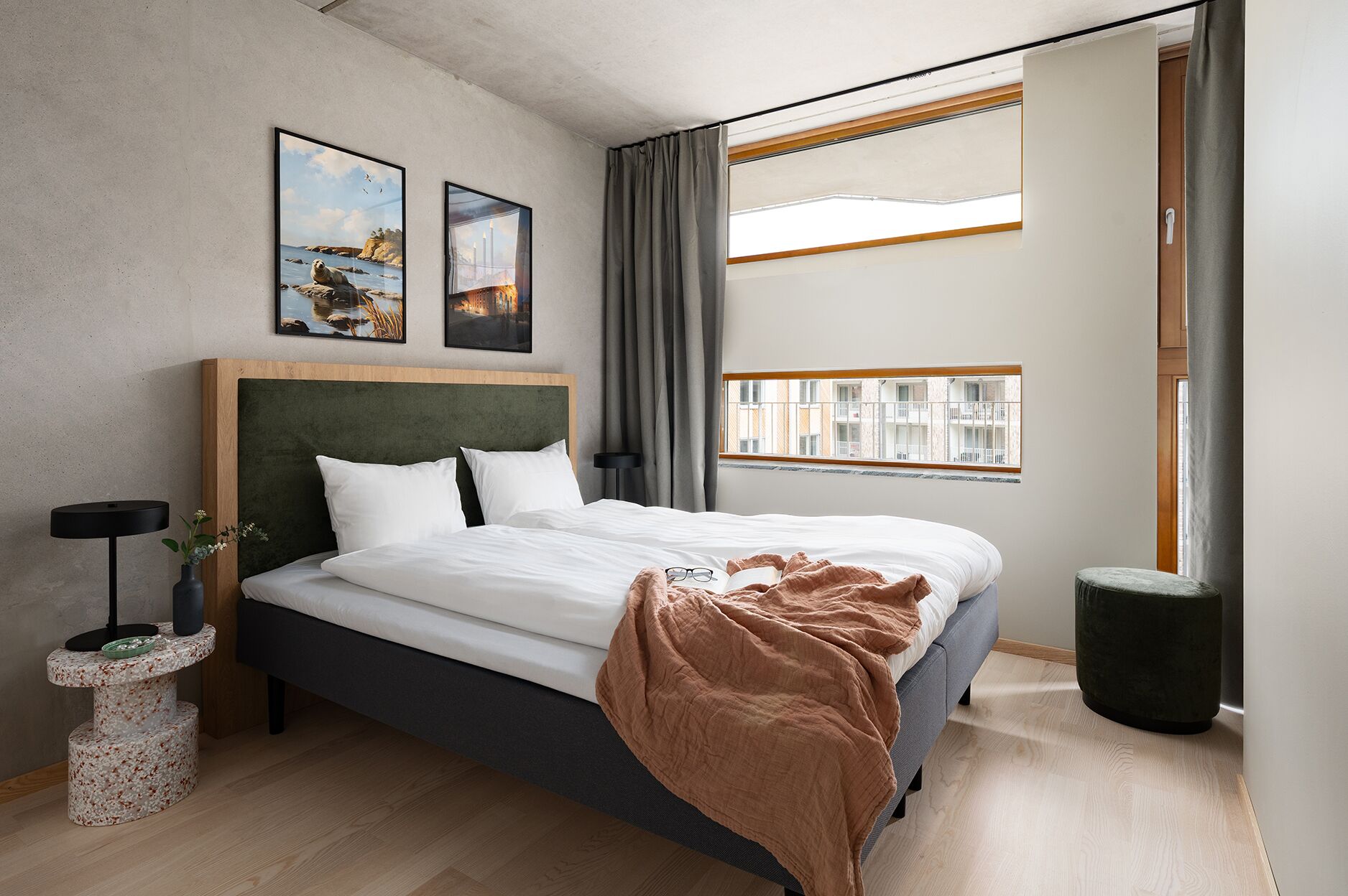 Un dormitorio moderno con una cama grande, un cabecero, una mesita de noche con un jarrón y una ventana con vistas a edificios.