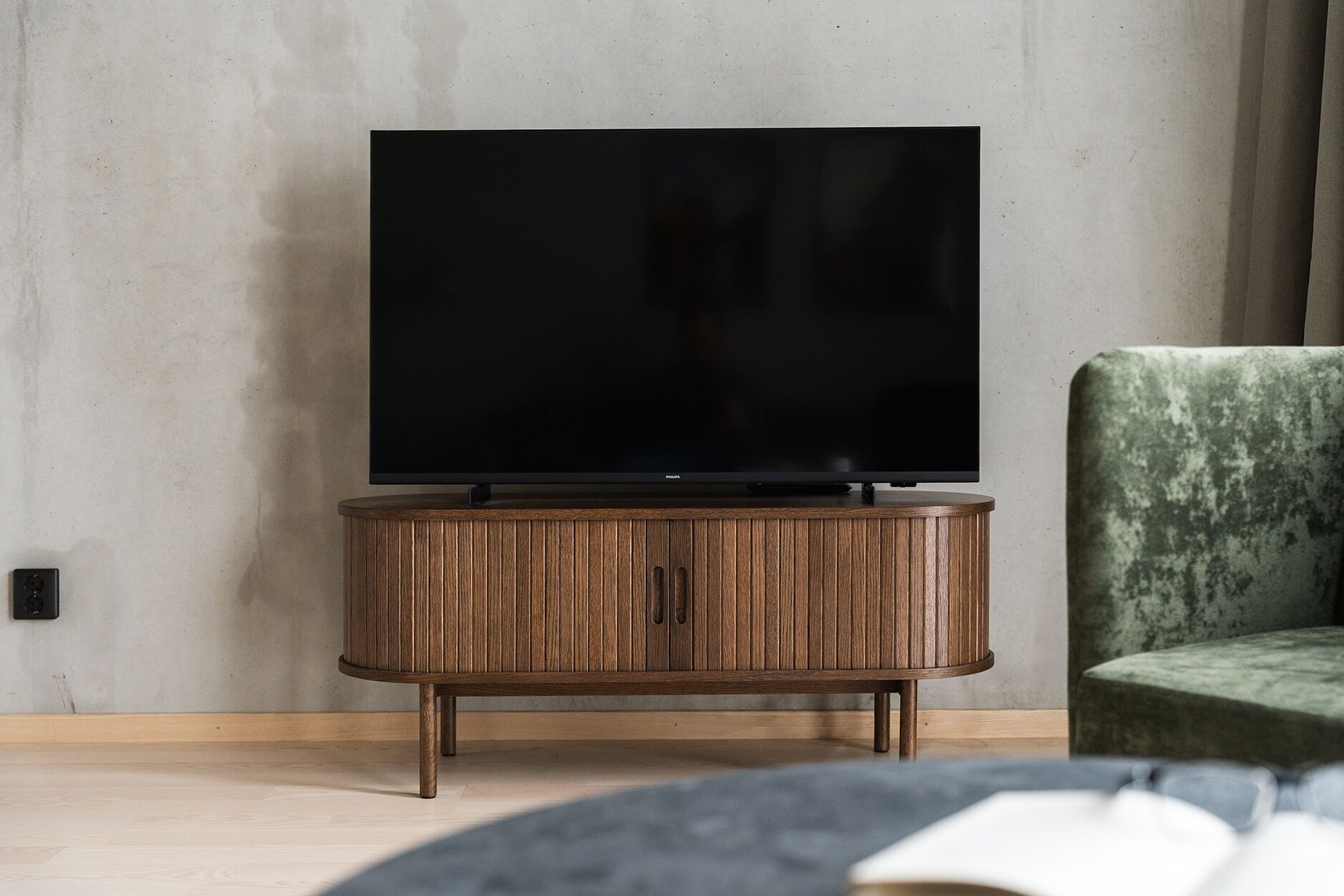 Un salon moderne avec un téléviseur à écran plat posé sur un meuble en bois, un fauteuil recouvert d’un tissu vert et un livre sur une table.