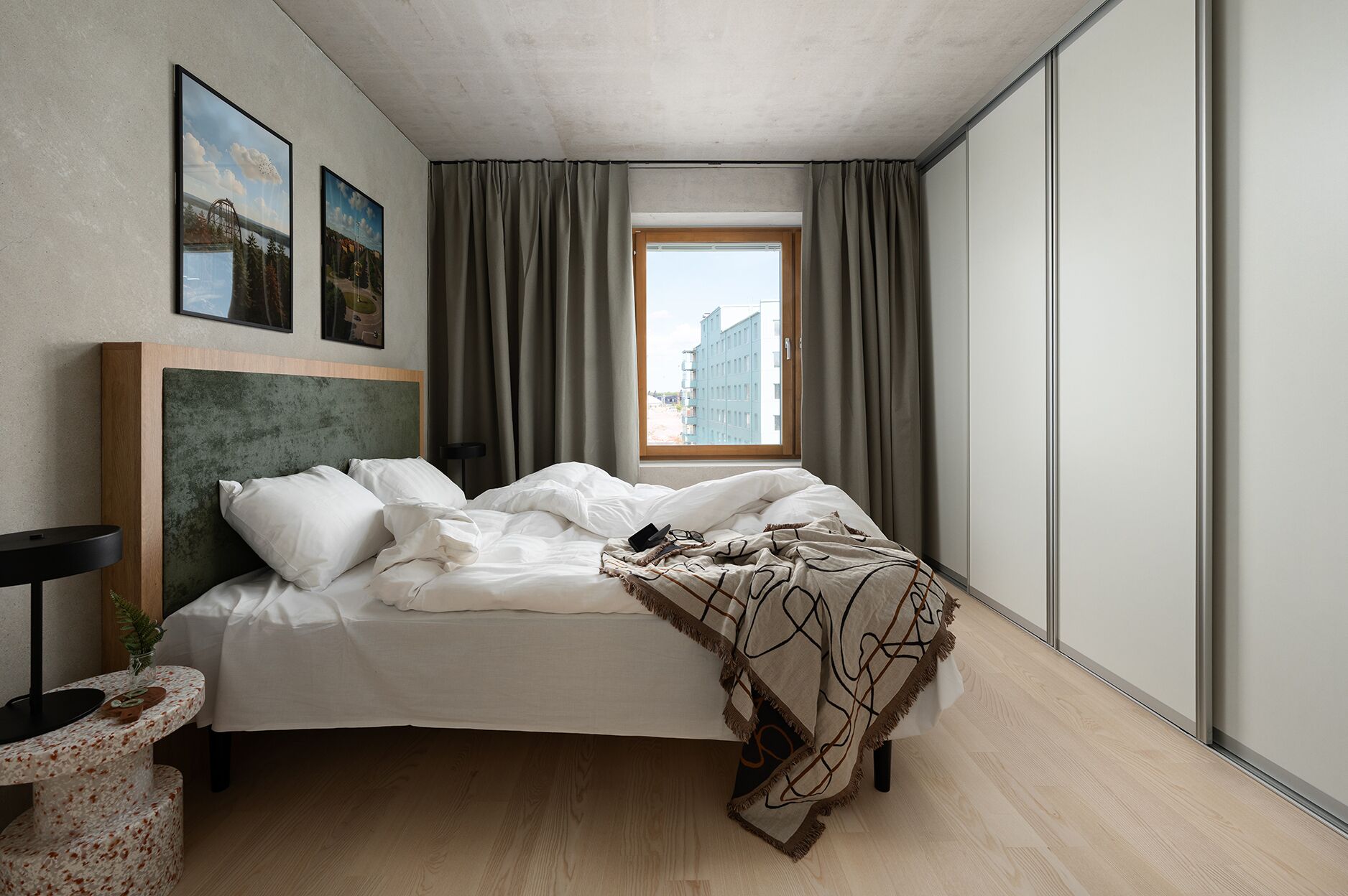 A modern bedroom with a large bed, a window with curtains, a headboard, and a small table with a plant.