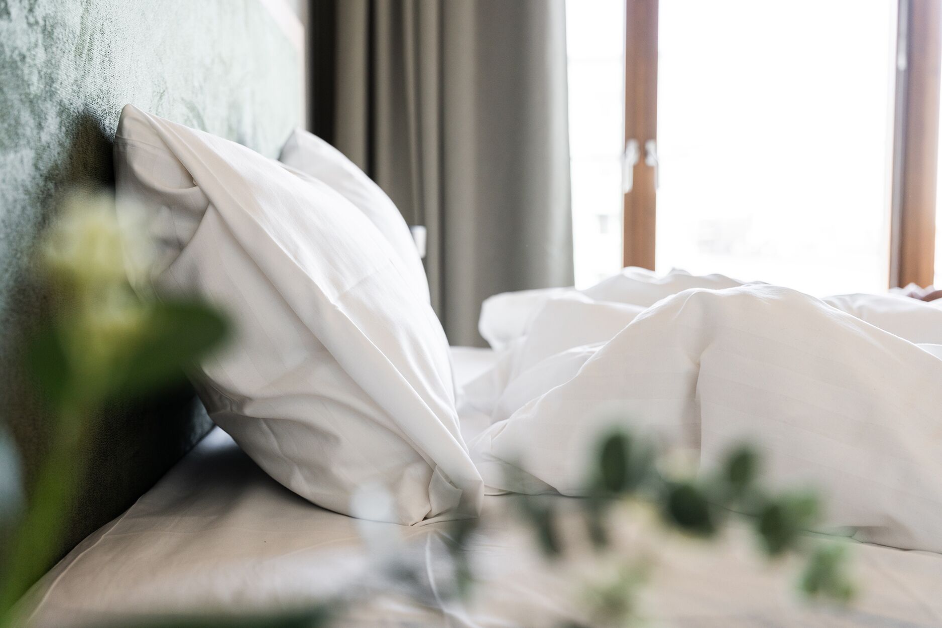 A bed with white bedding and pillows, a window with curtains, and a plant in the foreground.