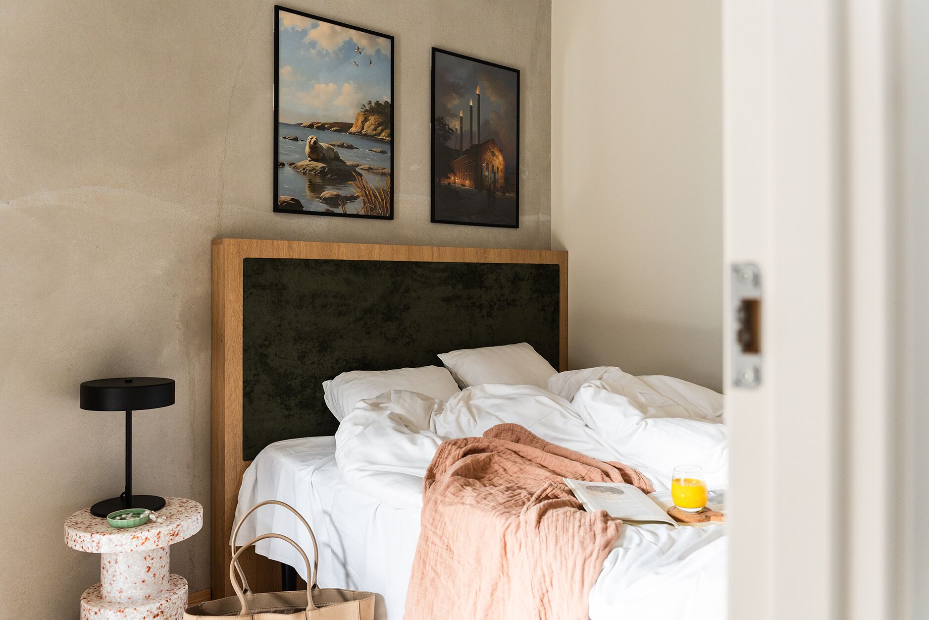 A neatly made bed with white linens, a pink throw blanket, and a glass of orange juice on the bedside table. There is a cat tree with a bowl and a green bowl on the floor beside the bed. Two framed pictures hang on the wall above the bed.