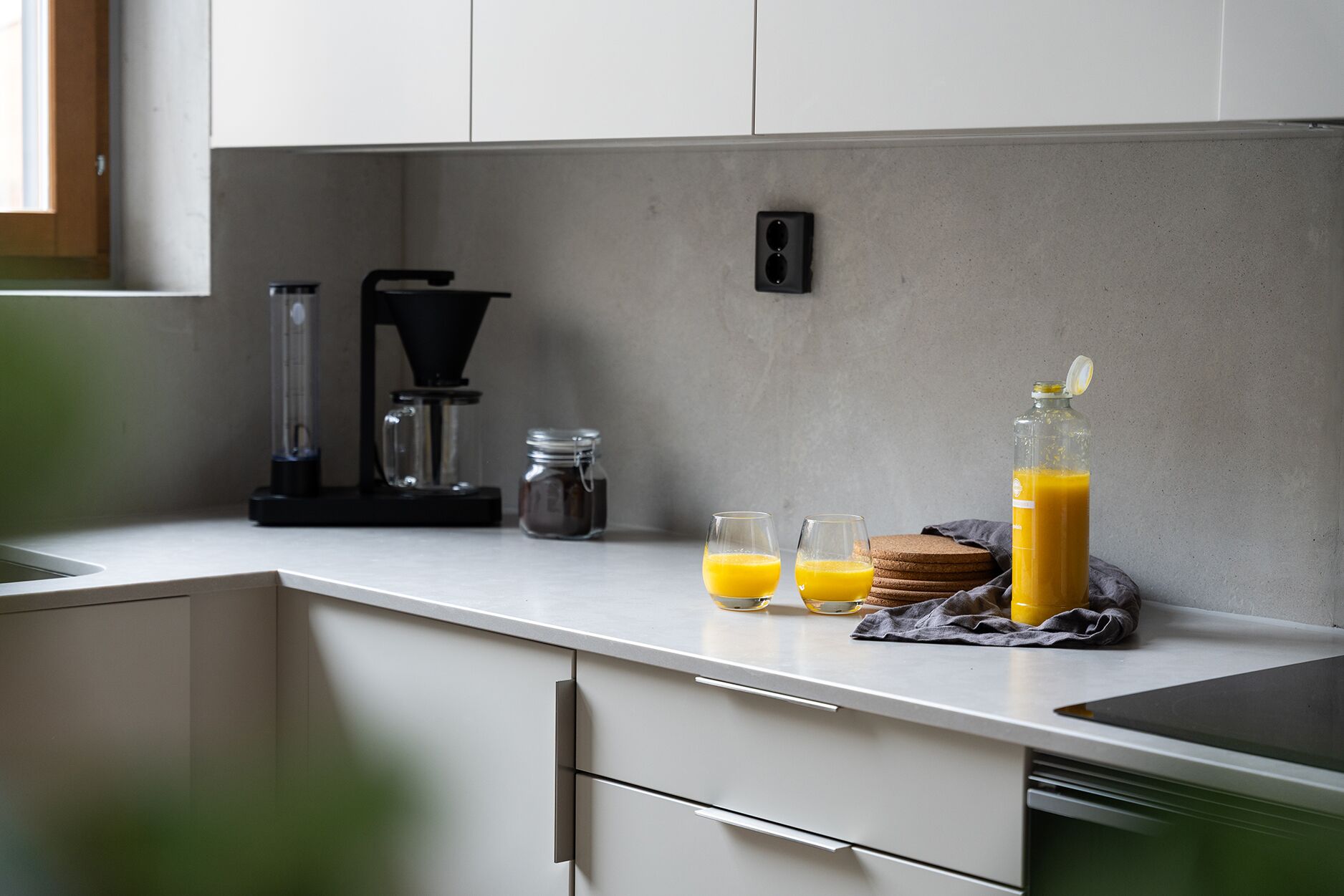 A kitchen with a white countertop, a coffee maker, a glass jar, two glasses of orange juice, a bottle of orange juice, and a cloth napkin.