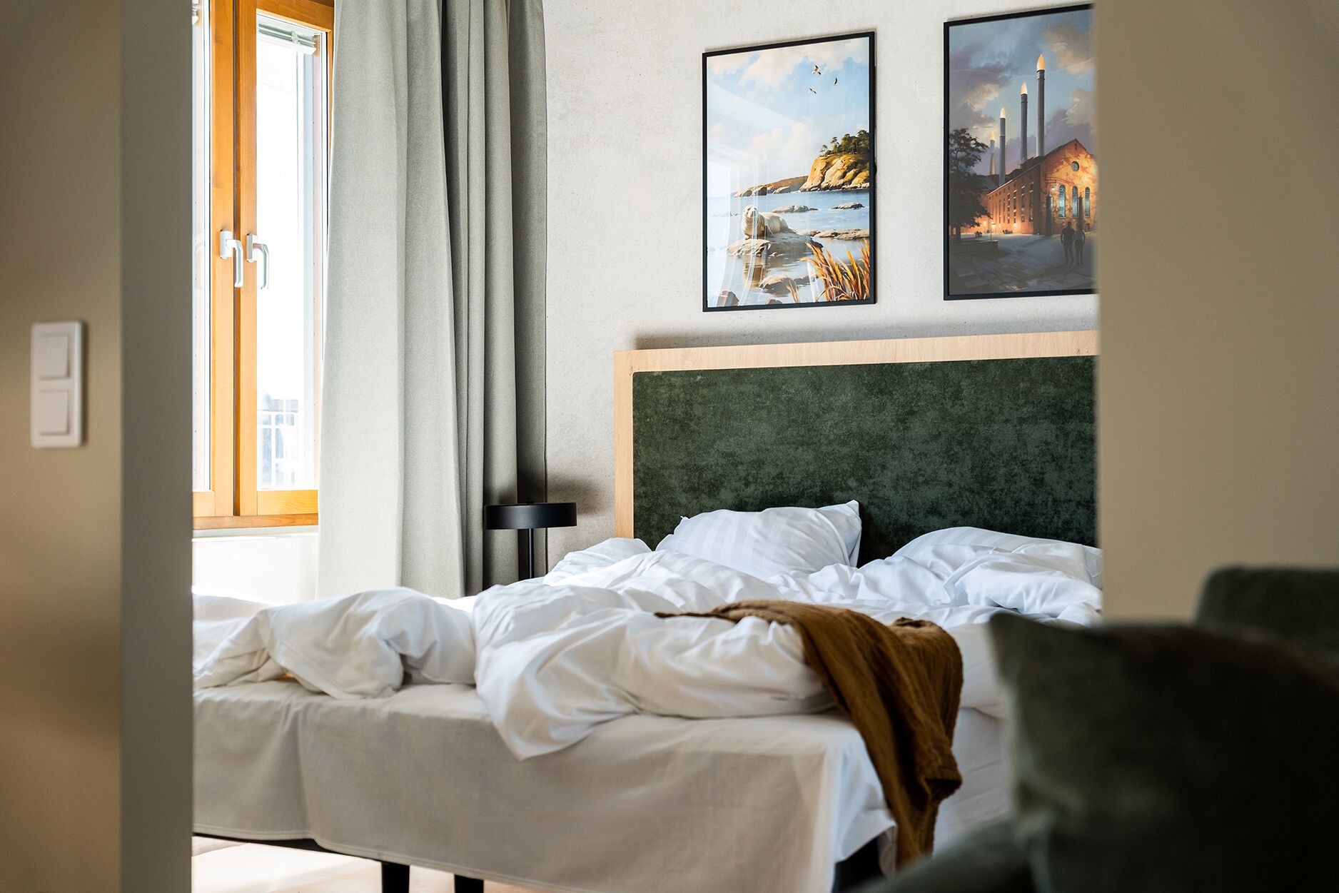 A hotel room with a bed, a headboard, a window with curtains, and two framed pictures on the wall.
