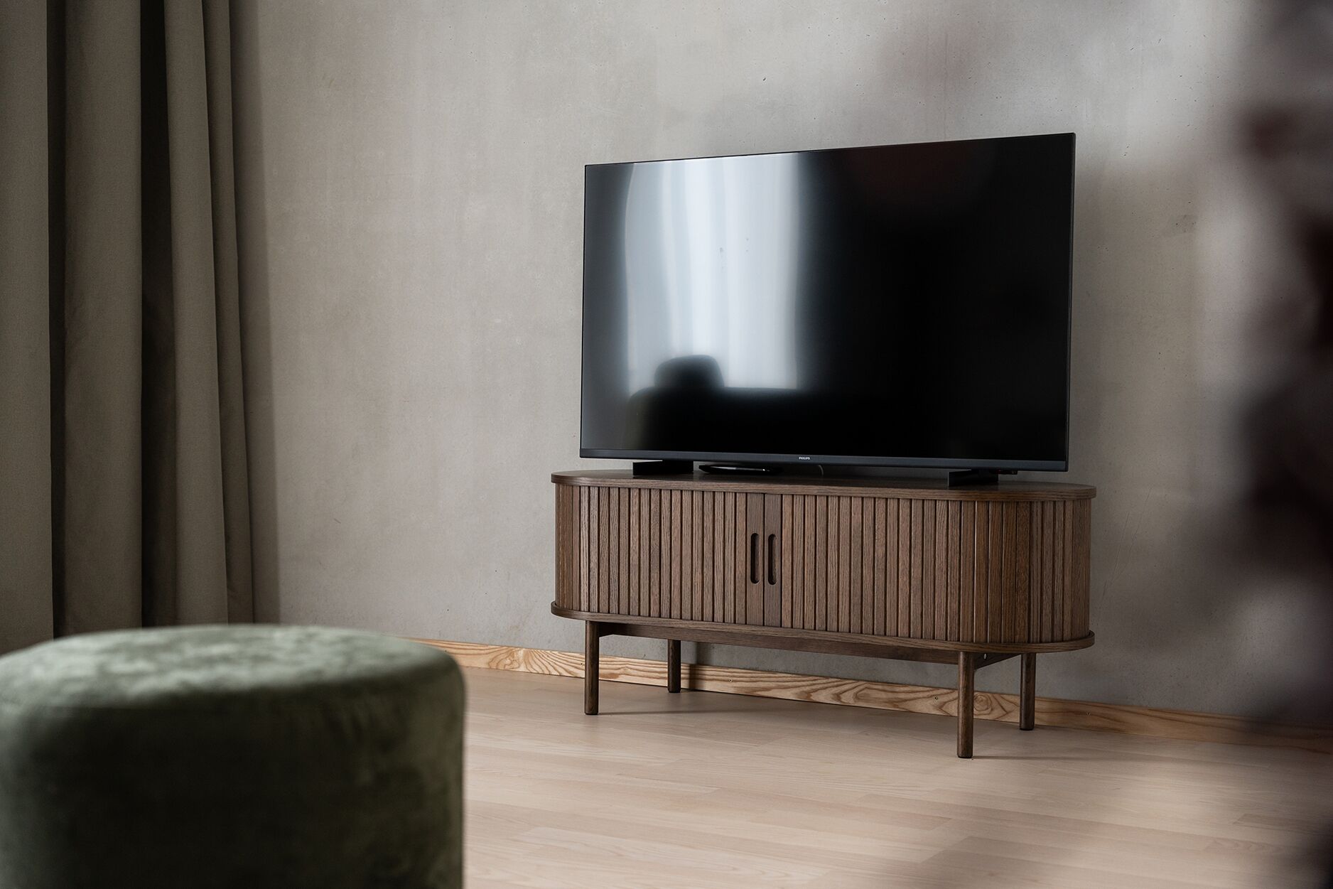 Een moderne woonkamer met een flatscreen televisie op een houten kast, een groene fauteuil en een effen muur.