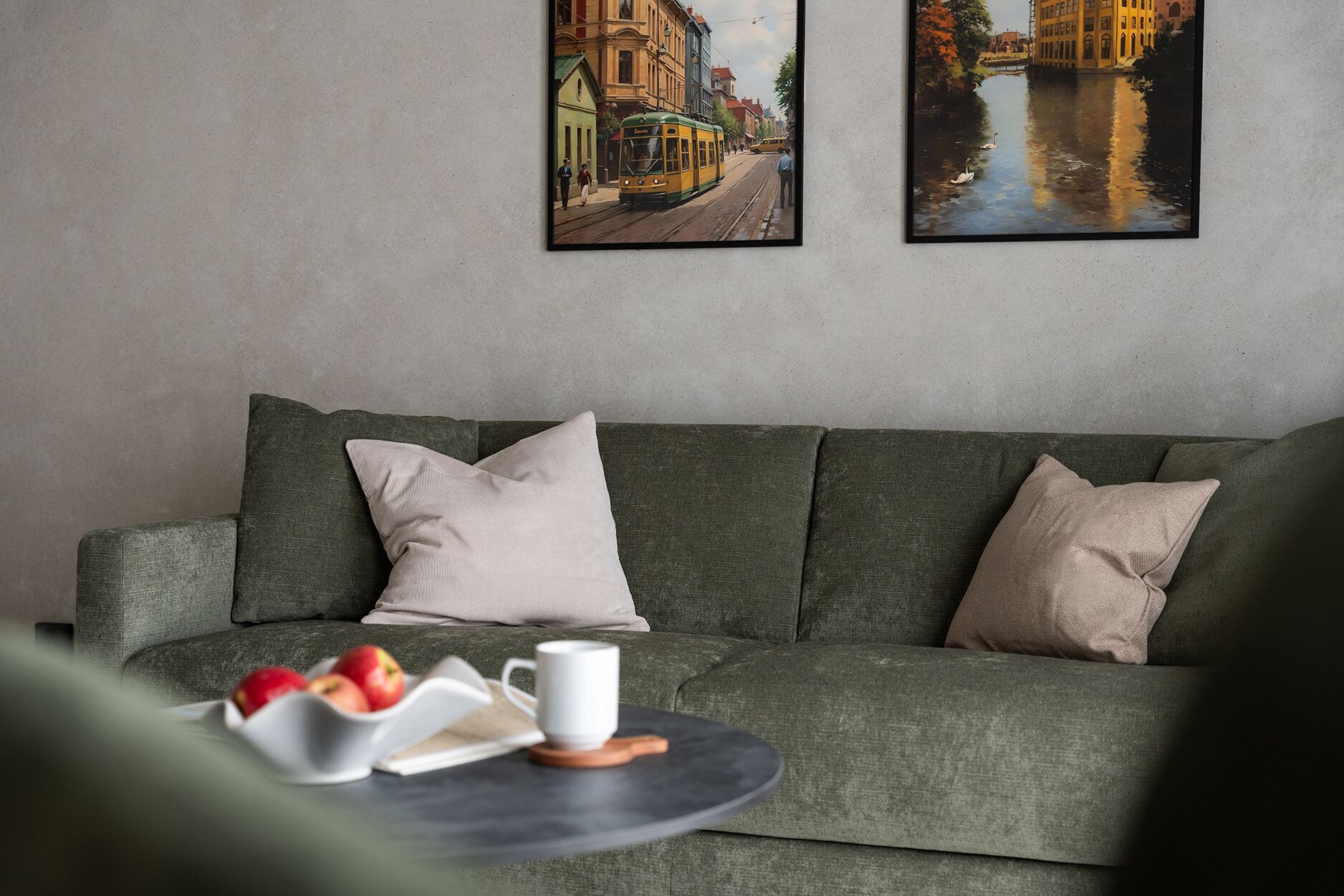 Una sala de estar moderna con un sofá verde, dos cojines, una mesita redonda con un libro, una taza y manzanas.