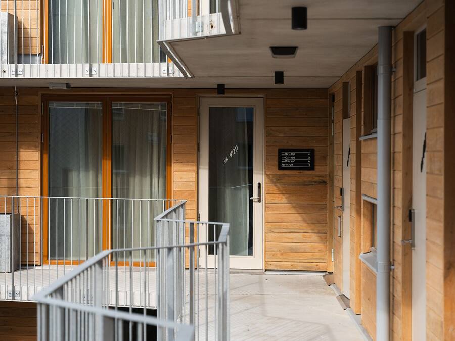 A modern building with a wooden facade, a glass door entrance, and a sign indicating room numbers and an elevator.