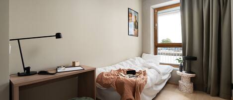 A bedroom with a bed, a desk, a chair, a window with curtains, and a small table with a plant.