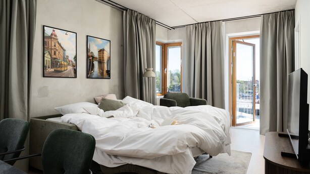 Une chambre d’hôtel avec un lit, deux chaises, une télévision et un balcon avec des rideaux.