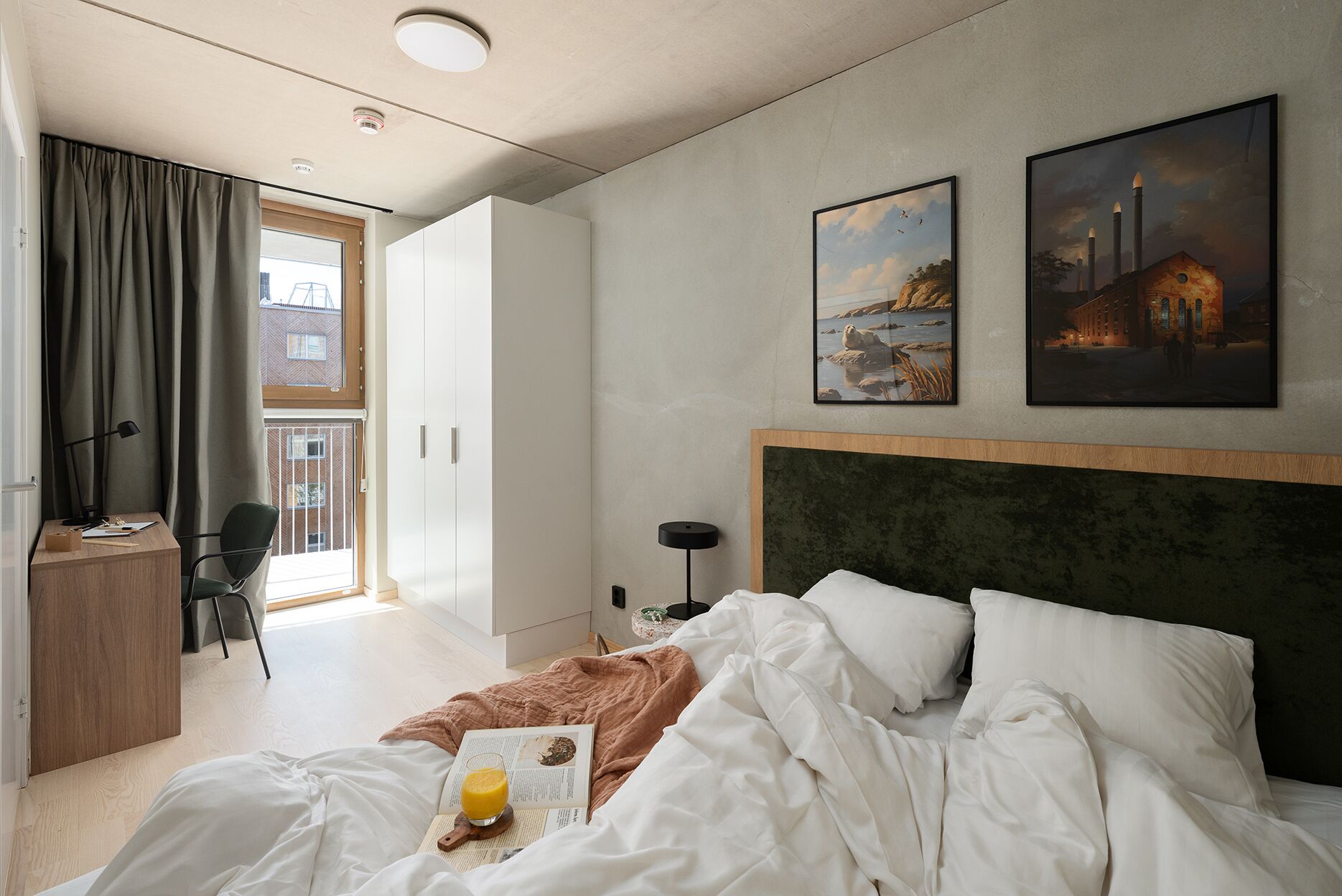 Habitación de hotel con cama, escritorio, silla y ventana con cortinas. Hay dos cuadros en la pared.