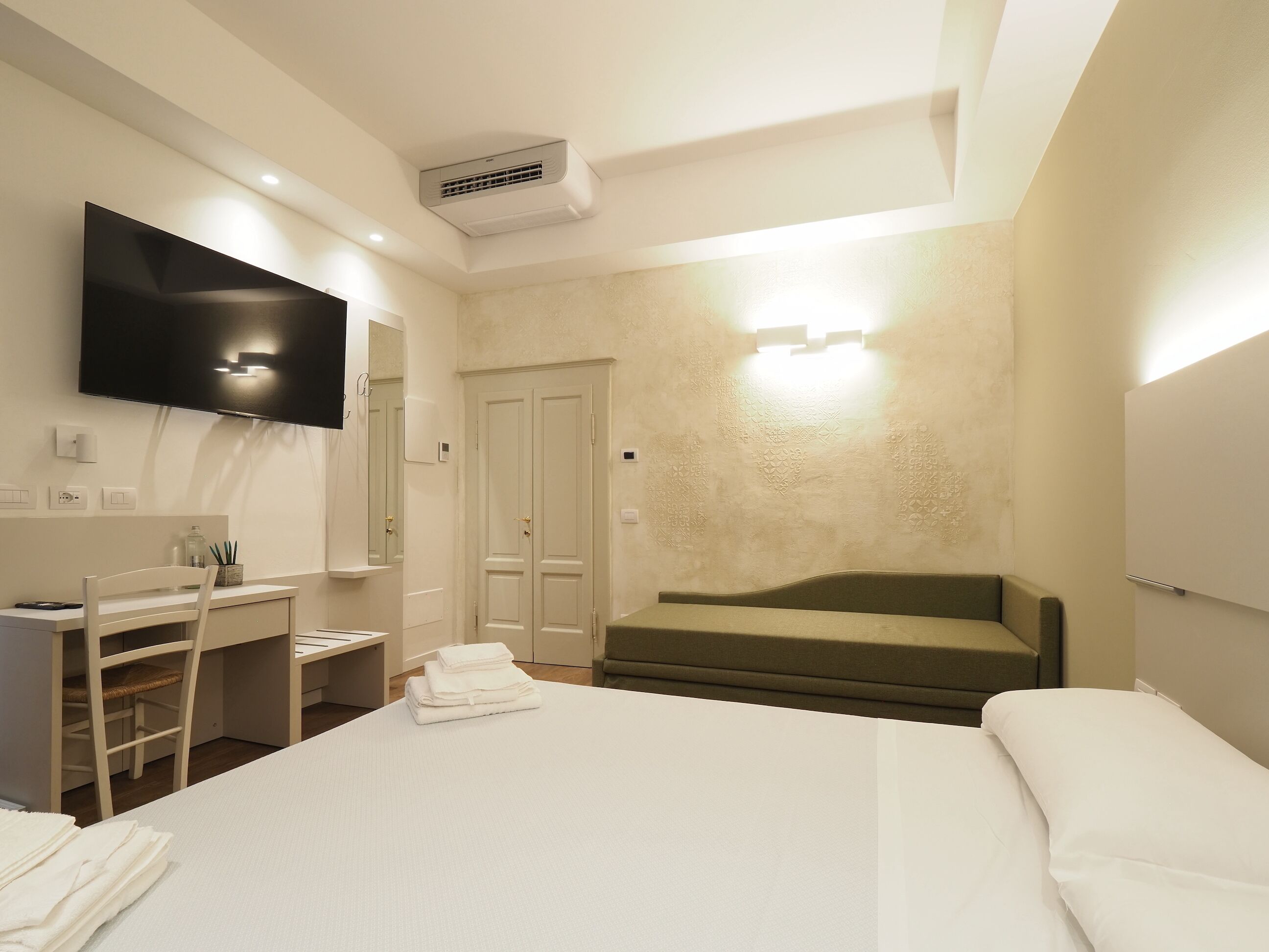 A hotel room with a bed, a desk with a chair, a television, a wardrobe, and a sofa.