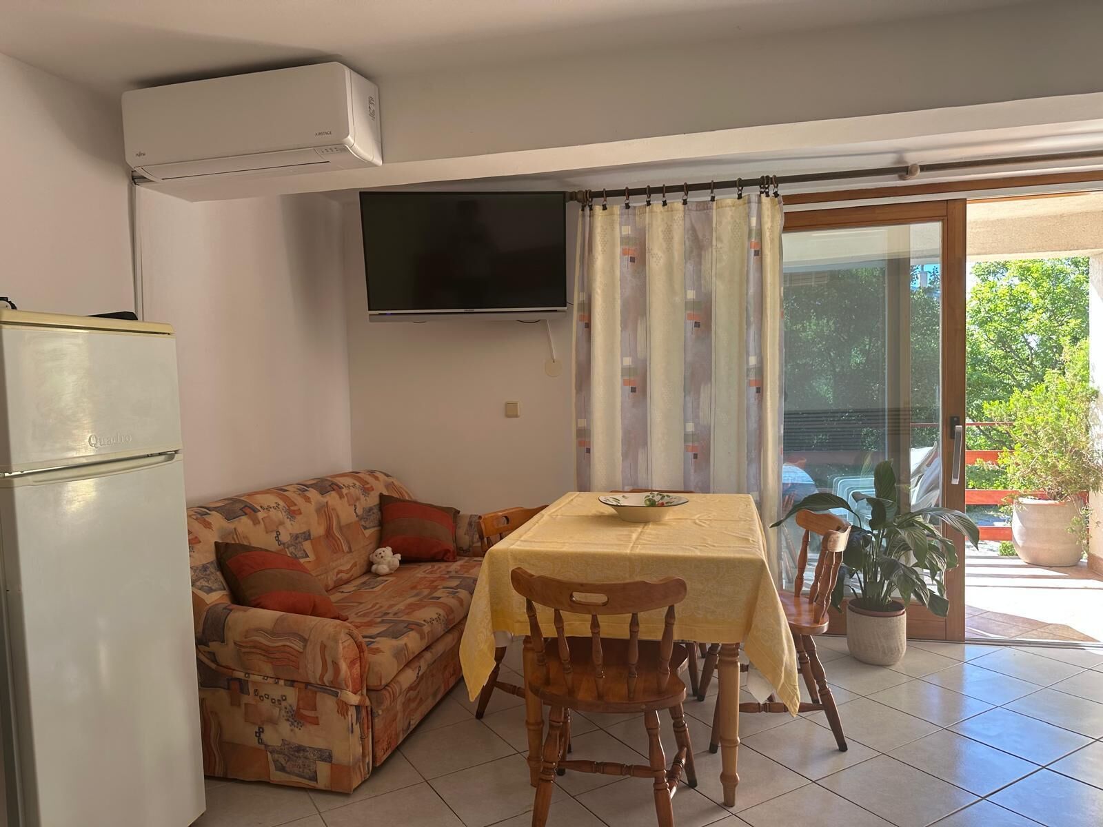 A living room with a sofa, a dining table with chairs, a television, and a balcony with potted plants.