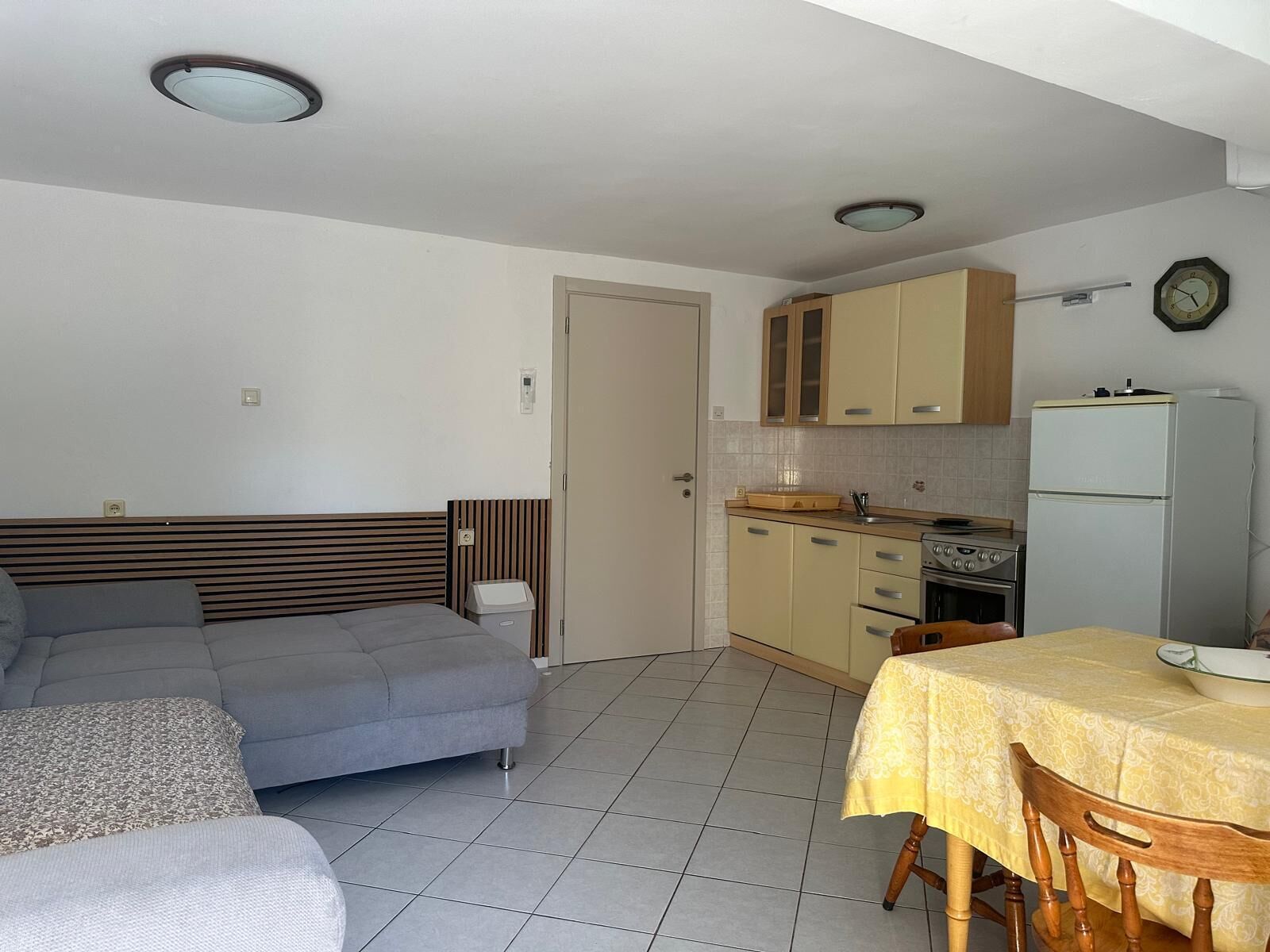 A compact living space with a sofa, a dining table, a kitchenette with a stove and refrigerator, and a door leading to another room.