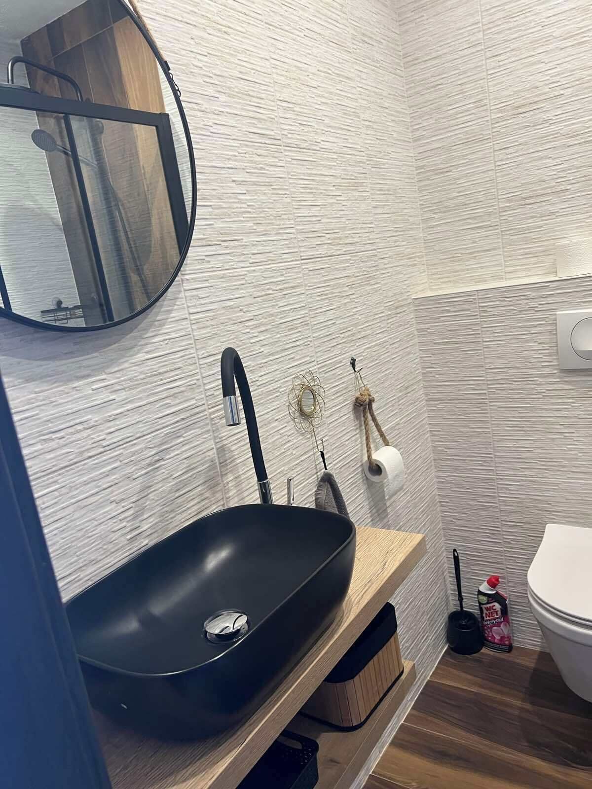 Un baño moderno con un lavabo negro, tocador de madera, un espejo redondo y un inodoro.
