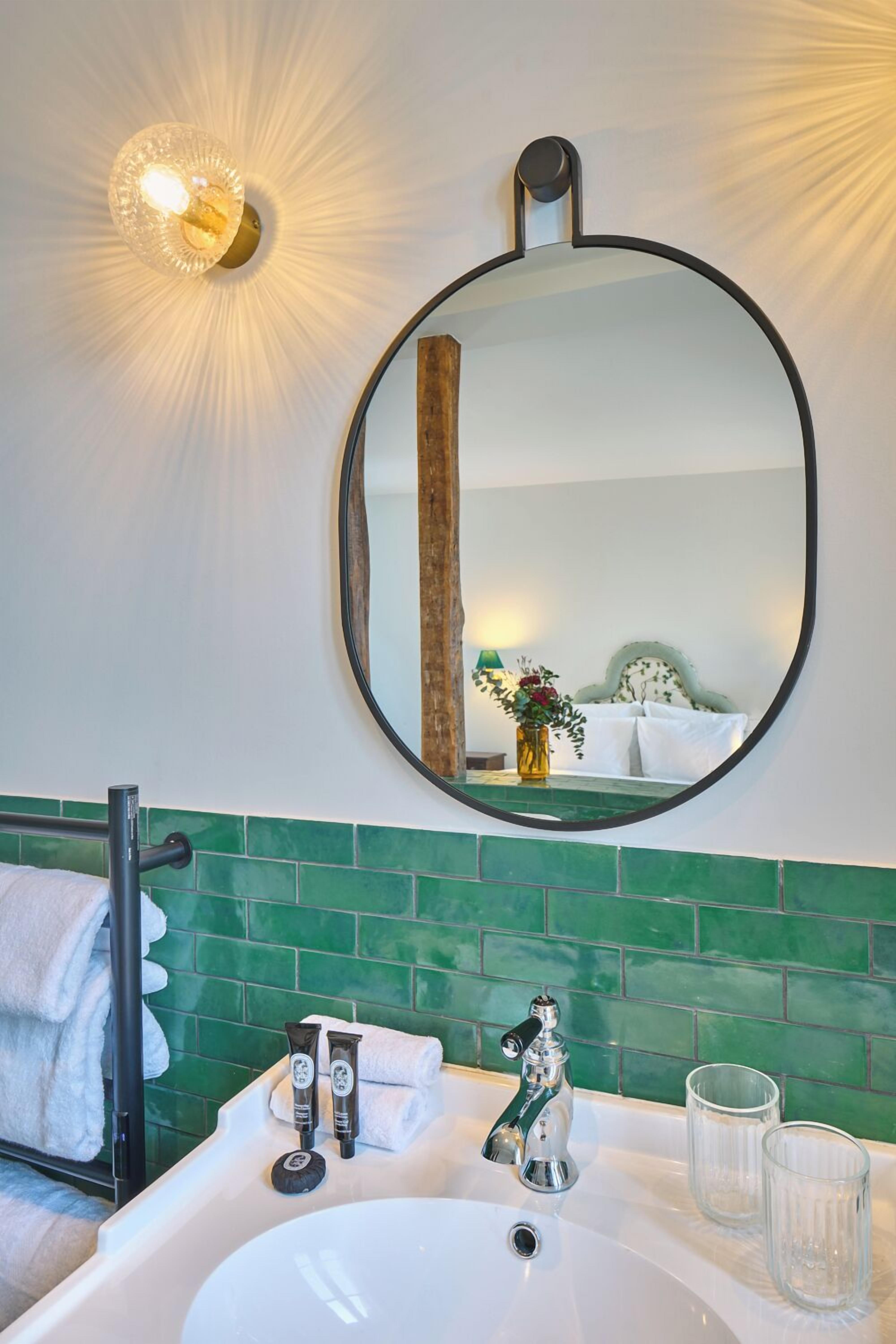 A bathroom with a round mirror, green tiled backsplash, a white sink, two toiletries, and a towel rack.