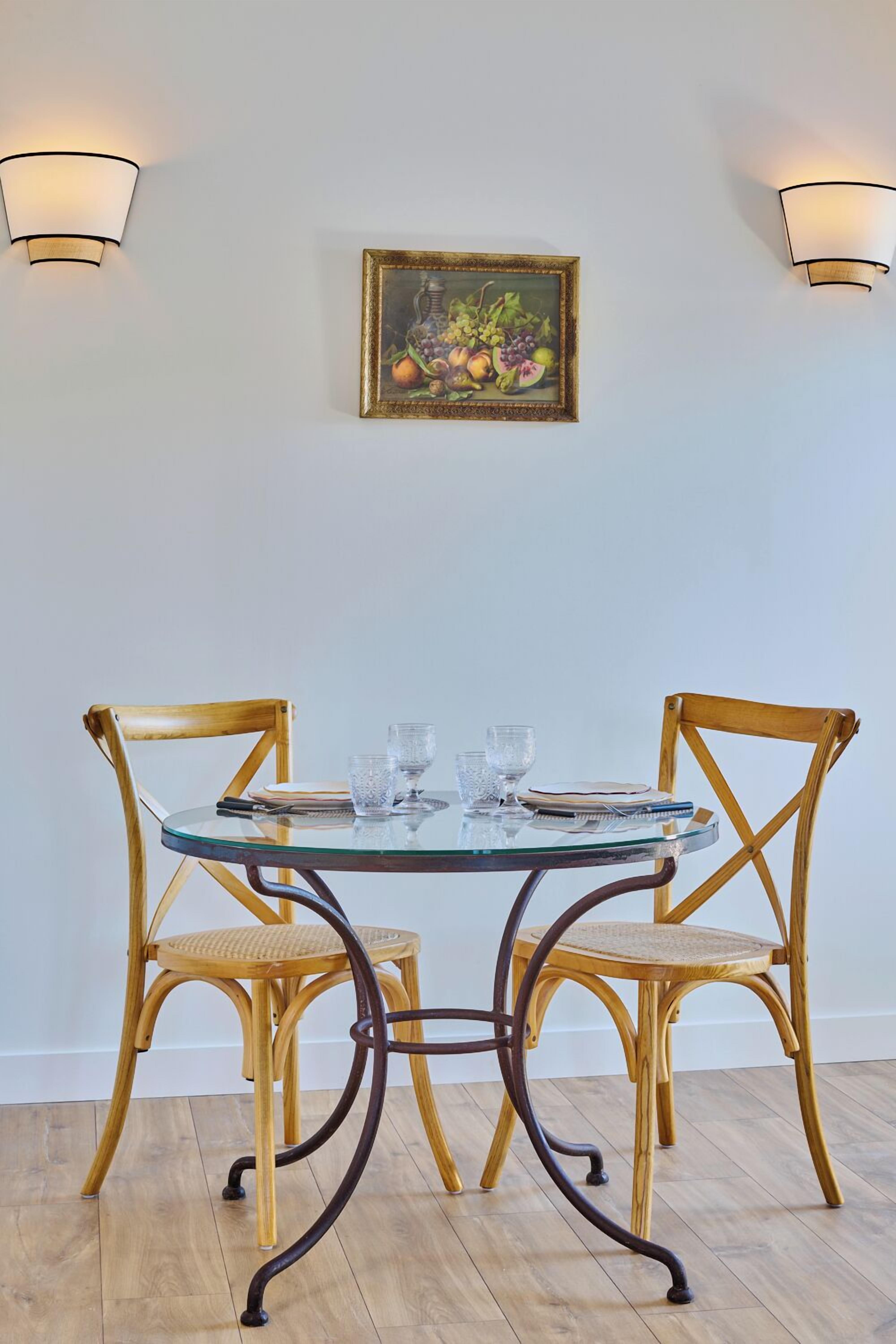 A dining table with two chairs, a glass top, and a framed painting on the wall.