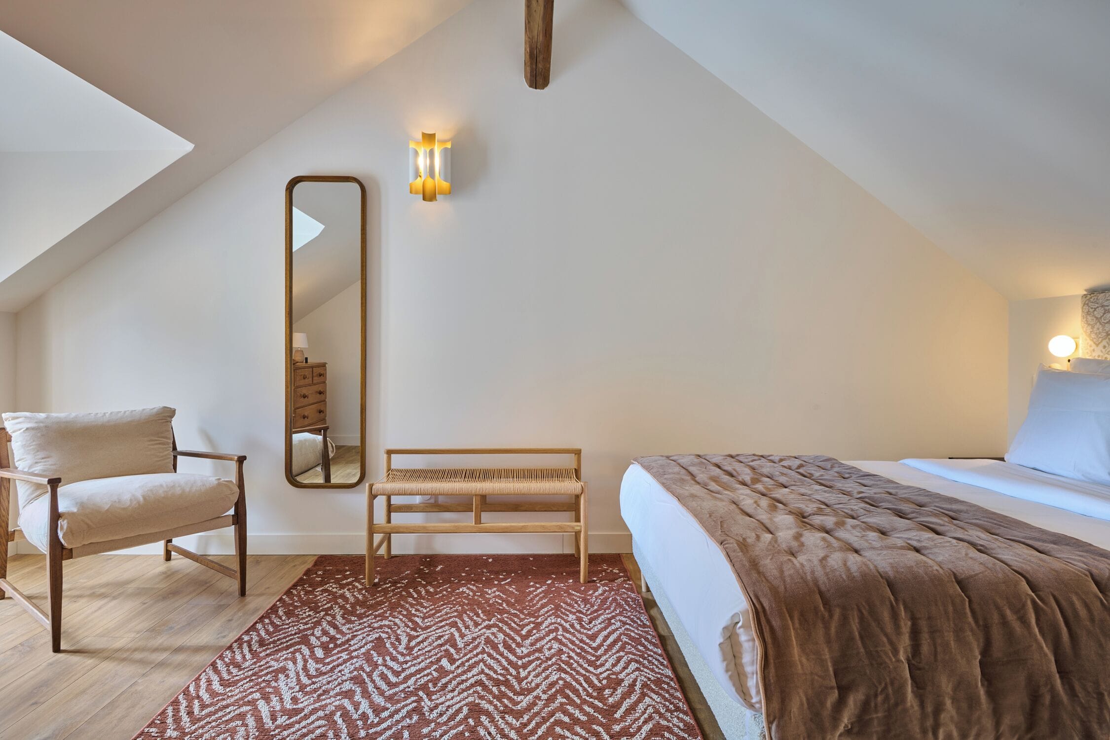 Une chambre à coucher comprenant un lit, une chaise, un banc, un miroir et un luminaire fixé au mur.