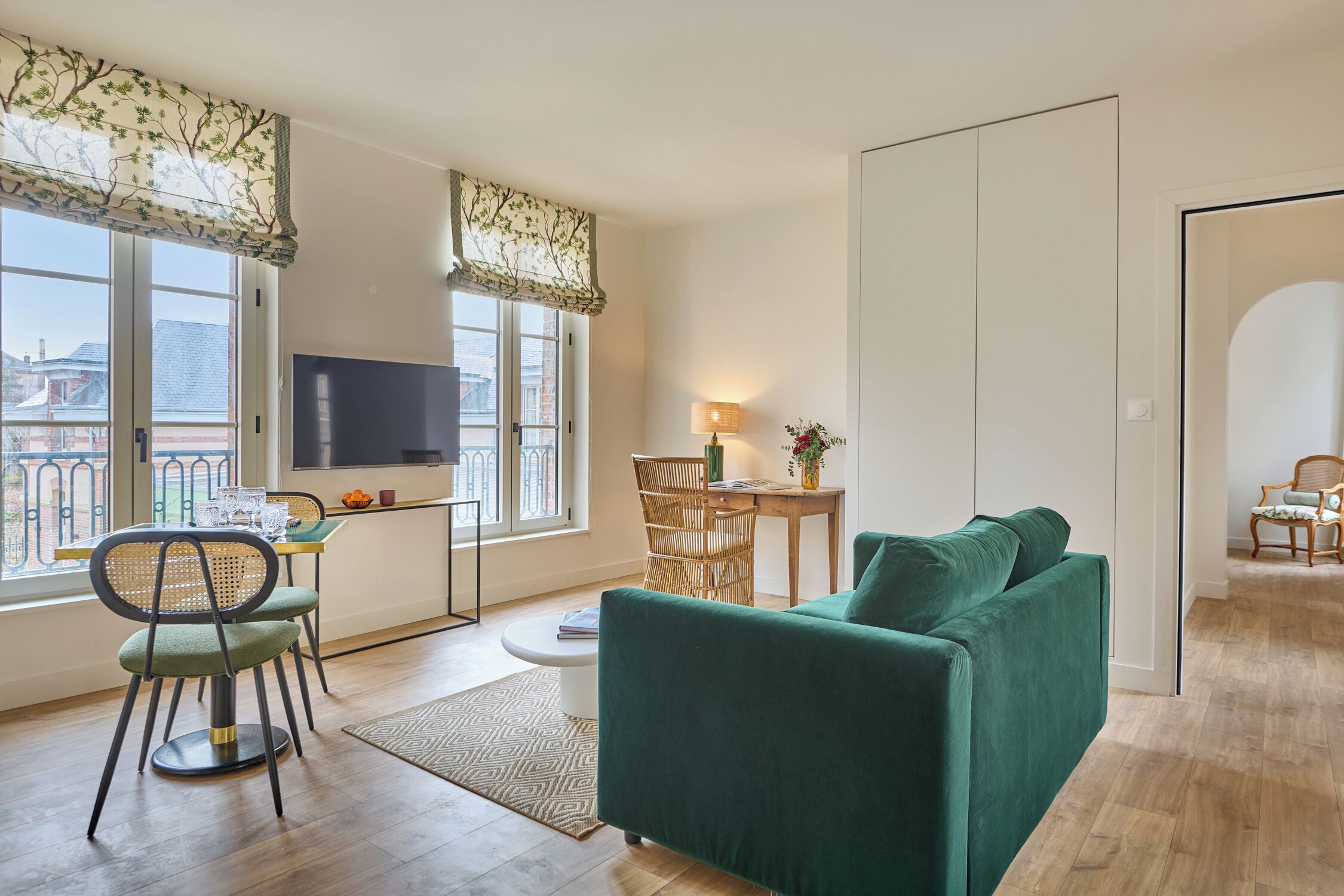 A modern living room with a green sofa, a dining table with chairs, a TV mounted on the wall, and large windows with curtains.