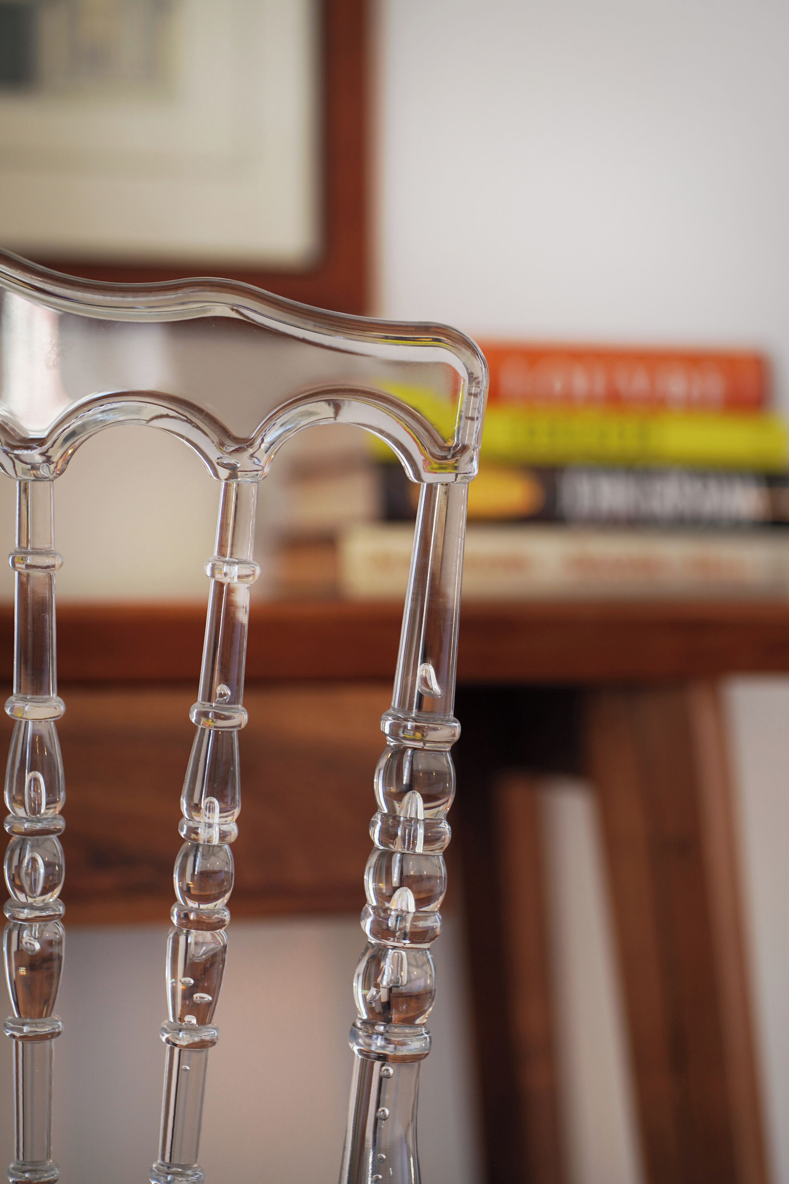 A clear, transparent chair with a curved backrest and armrests, positioned in front of a wooden desk with books.