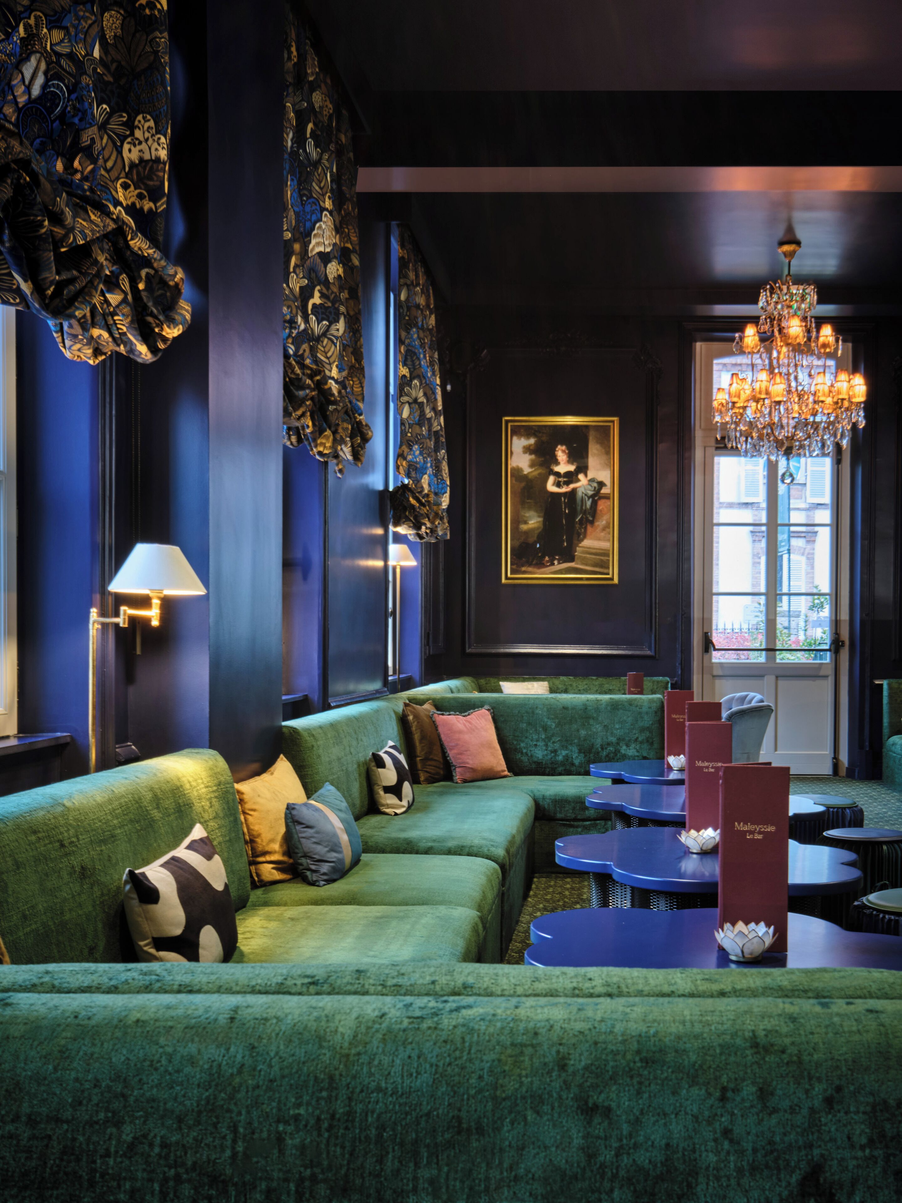 A modern lounge with a green sofa, patterned curtains, a chandelier, and a framed portrait on the wall.