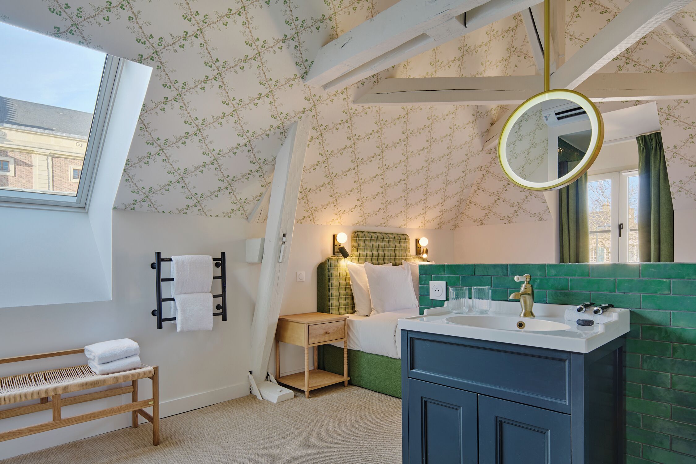 Un dormitorio con tragaluz, una mesita de noche, un banco, cabecero verde, cama blanca, espejo redondo, lavabo y pared con azulejos verdes.