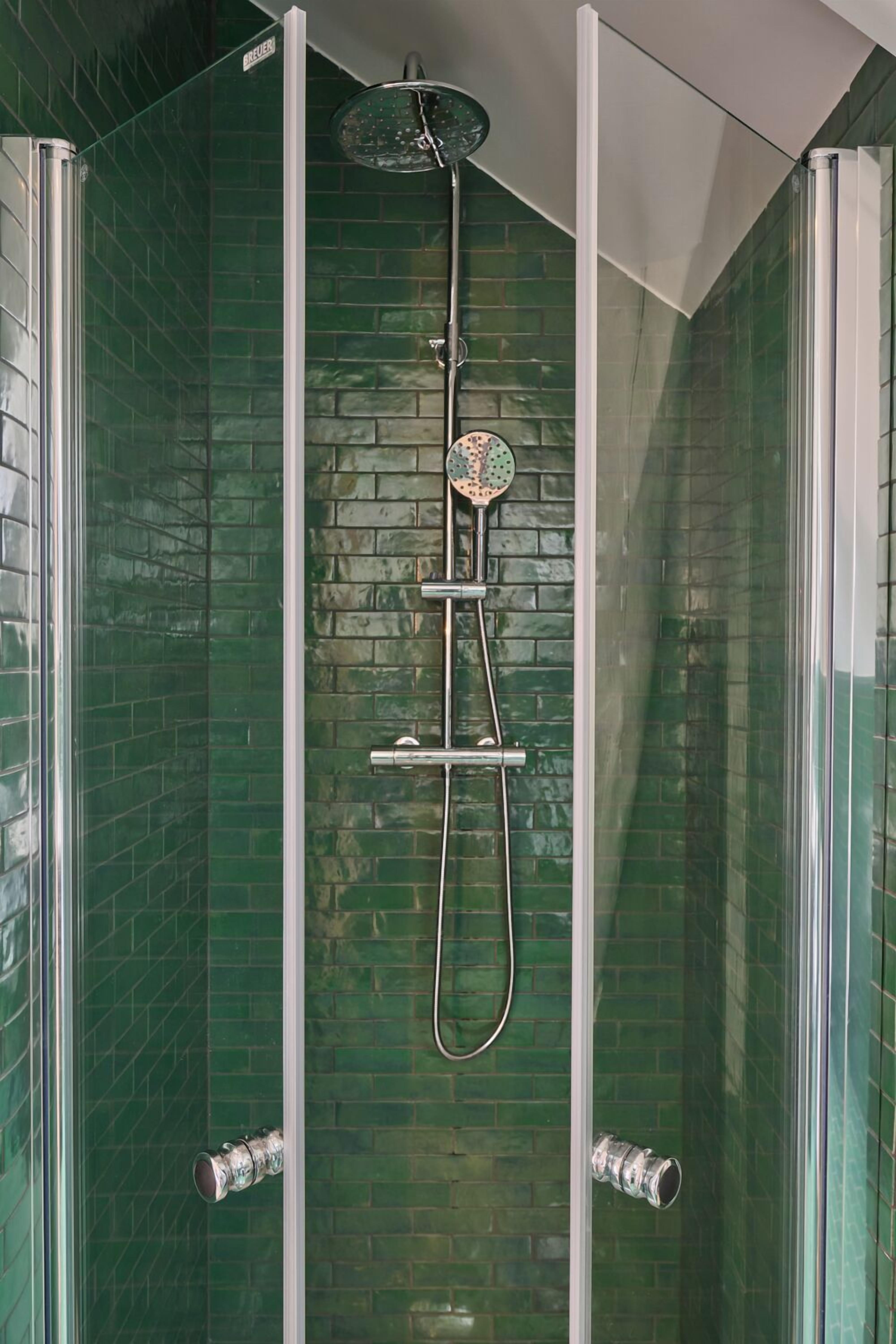 Ducha envuelta en vidrio con cabezal de ducha de lluvia y otro manual, frente a una pared con azulejos verdes.