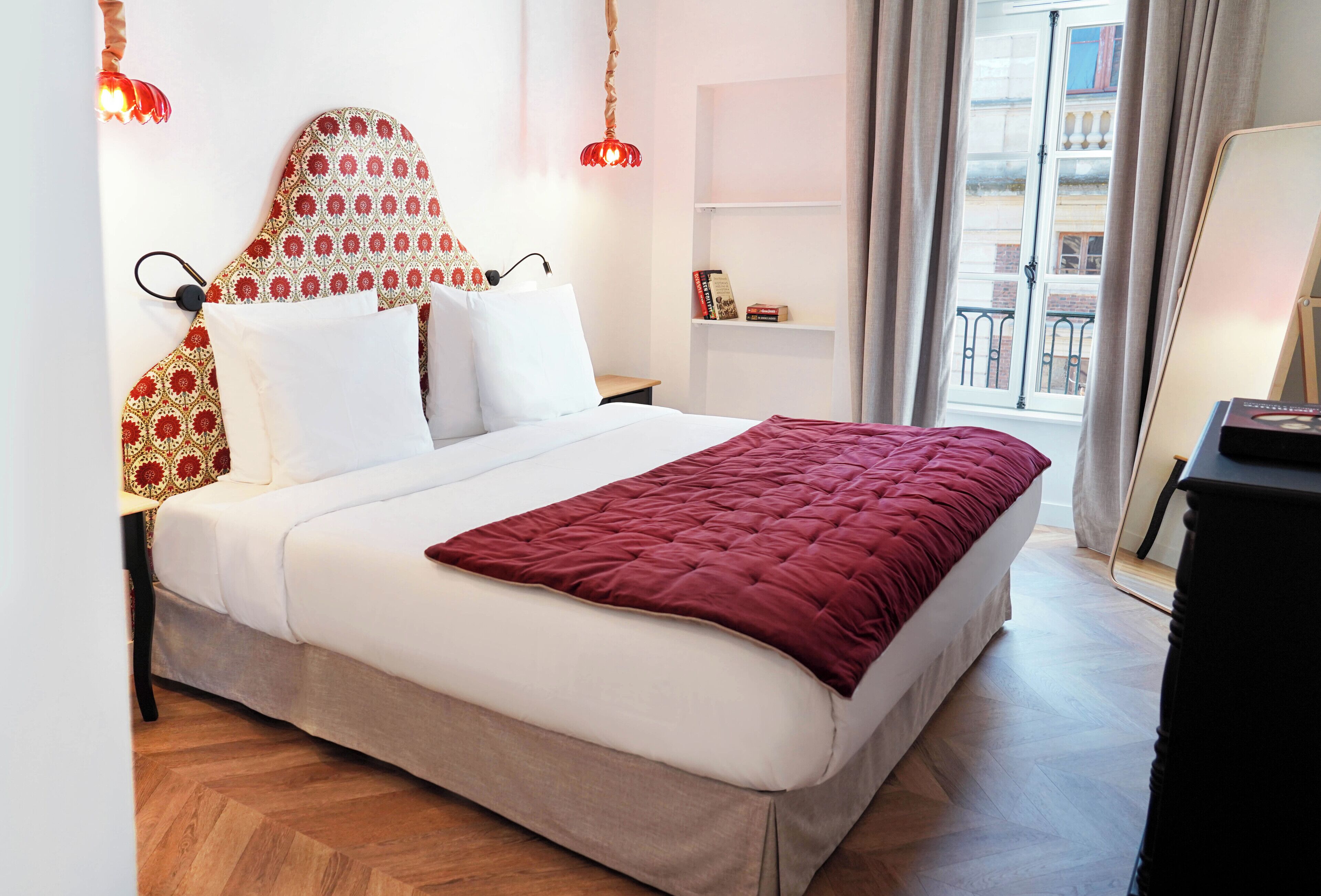 Una cama bien tendida con una colcha roja, un cabecero estampado y dos lámparas de noche.