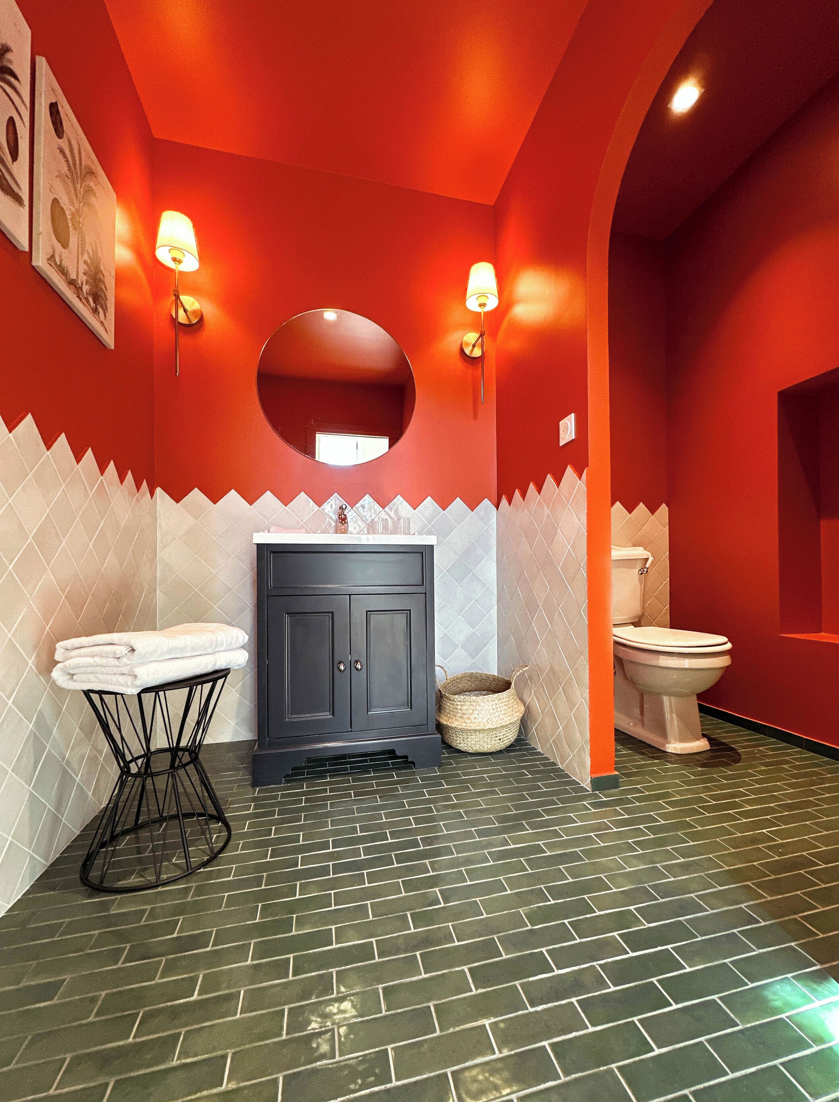 Ein Badezimmer mit roter Wand, einem weißen Waschtisch, einem schwarzen Schrank, einem runden Spiegel, einer weißen Toilette und einem schwarzen Boden.