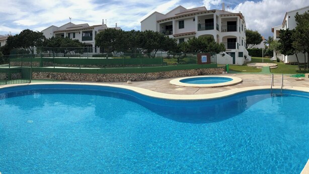 A swimming pool with a curved edge, surrounded by a stone wall and green fencing. - APARTMENT IN CALA'N FORCAT (Los Delfines)