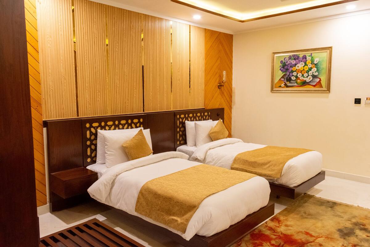 A hotel room with two beds, wooden headboards, a wooden panel wall, a painting, and a small nightstand.