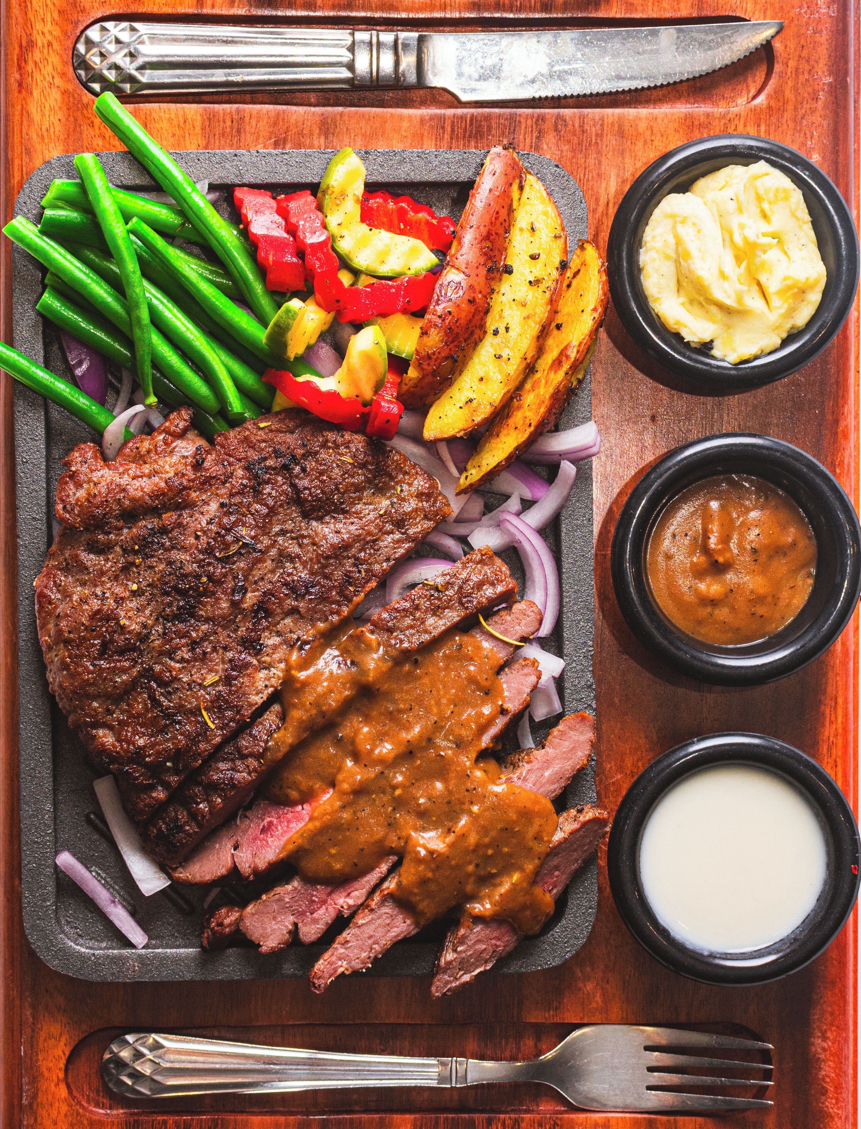Un steak nappé de sauce, accompagné de haricots verts, de pommes de terre rôties et de poivrons rouges, le tout servi sur un plateau en bois.