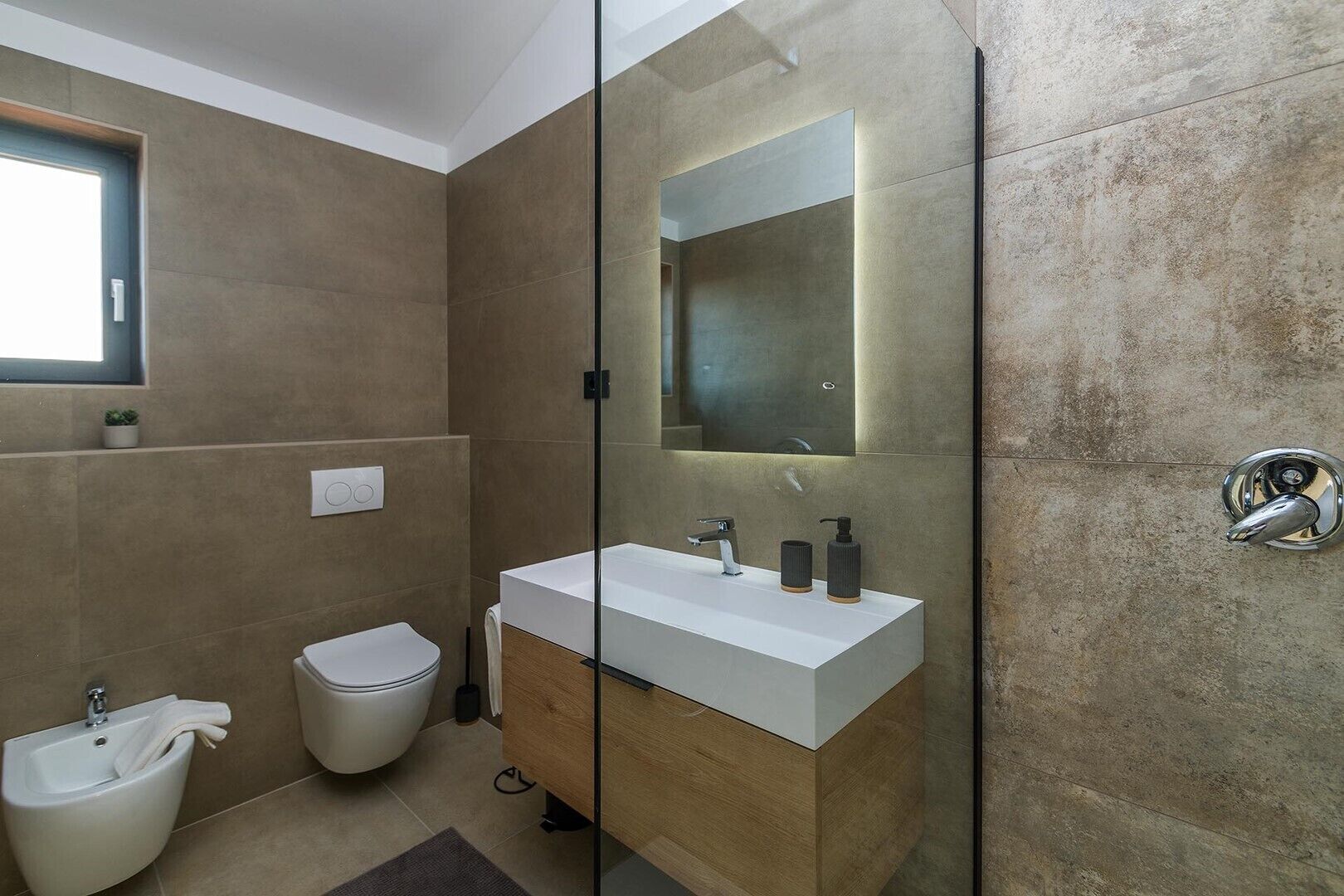 Une salle de bain moderne dotée d’une douche à parois de verre, d’un lavabo blanc avec un meuble-vasque en bois, de toilettes et d’un bidet.