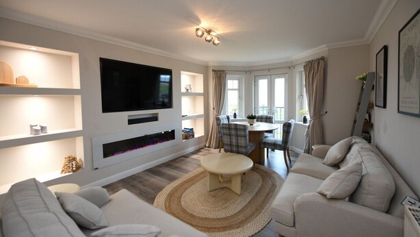 A modern living room with a flat-screen TV, built-in shelves, a dining area with plaid chairs, and a large window with curtains. - Barassie Bay View apartment (Troon)