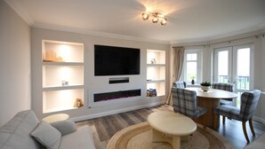 A modern living room with a flat-screen TV, built-in shelves, a fireplace, and a dining area with plaid chairs. - Barassie Bay View apartment (Troon)