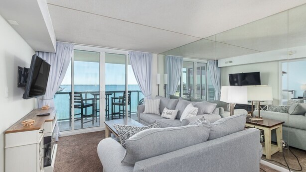 A modern living room with a sofa, a coffee table, and a TV. There is a balcony with a view of the sea. - Sunset Selfies & Seaside Memories | Bayfront OCMD Retreat (Ocean City)