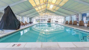 An indoor swimming pool with a covered roof, clear glass walls, and wooden lounge chairs around the pool. - Sunset Selfies & Seaside Memories | Bayfront OCMD Retreat (Ocean City)