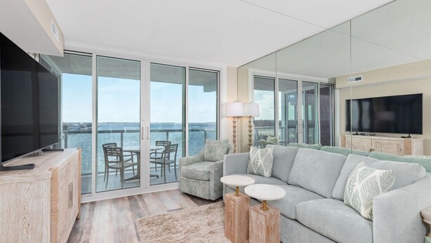 A modern living room with a large TV, a grey sofa, and a balcony with a view of the sea. - Crab Feasts, Bay Breezes & Boardwalk Nights | Summer Starts Here (Ocean City)