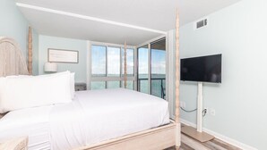 A bedroom with a large bed, a TV mounted on the wall, and a view of the sea through large windows. - Crab Feasts, Bay Breezes & Boardwalk Nights | Summer Starts Here (Ocean City)