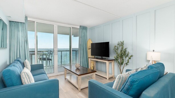 Un salon moderne doté d’une grande porte-fenêtre coulissante donnant sur un balcon avec vue sur la mer. On y trouve un téléviseur posé sur un meuble en bois et une petite table sur laquelle est placé un objet décoratif.