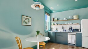 Een keuken met teal muren, een witte koelkast, een magnetron en een spoelbak. Er is een raam met zonwering, een witte tafel met stoelen en een plant op de tafel.