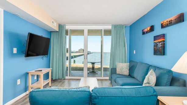 Una habitación con pared azul, un televisor de pantalla plana, un sofá, una mesa y vista al mar a través de una puerta de cristal.