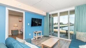 Una sala de estar moderna con pared azul, televisor de pantalla plana, sofá, mesa de centro y puerta corrediza de vidrio que da a un balcón con vista al agua.
