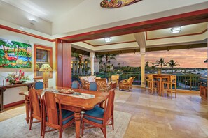 A spacious dining area with a wooden table and chairs, a view of the sunset, and a balcony with wicker furniture. - 🌴Aloha Moana Penthouse  | Elevator | Ocean View | Beach Access🌴 (Waikoloa)