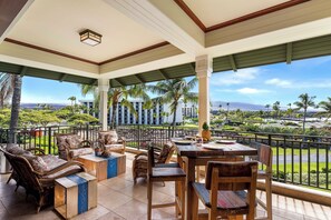 A covered outdoor seating area with wicker furniture and a wooden table, overlooking a lush landscape and hotel buildings. - 🌴Aloha Moana Penthouse  | Elevator | Ocean View | Beach Access🌴 (Waikoloa)