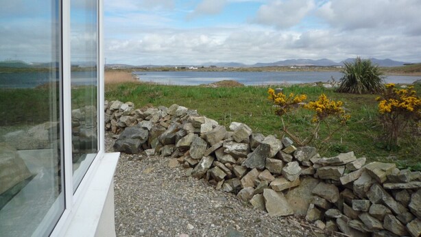 A view from a window overlooking a lake with a stone wall and yellow flowers in the foreground.