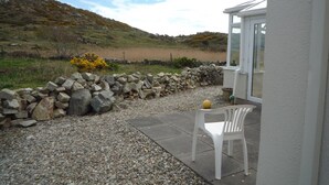 A patio area with a white plastic chair, a stone wall, and a view of a grassy hill.