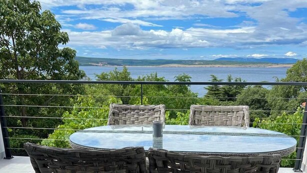 A round glass table with wicker chairs on a balcony overlooking a body of water and distant land. - Apartman Suzzy 2 (Dramalj)