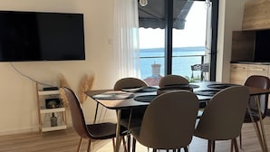 A dining area with a table and chairs, a flat-screen TV mounted on the wall, and a view of the sea through large windows. - Apartman Suzzy 2 (Dramalj)